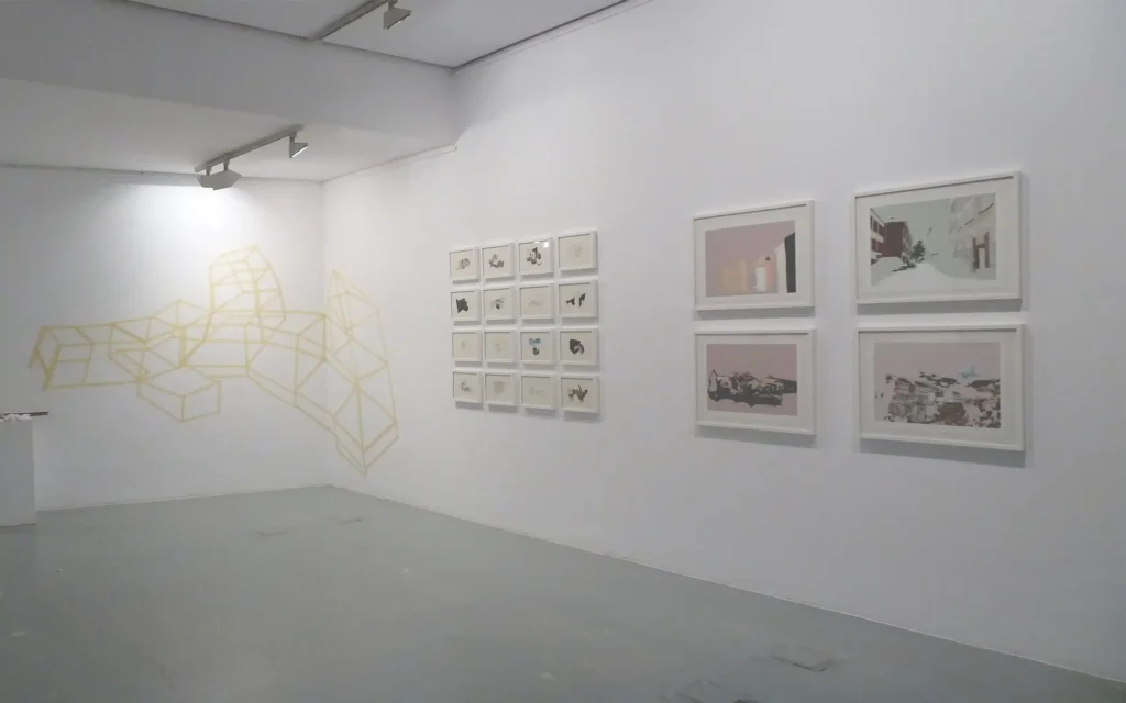 Sala de exposición encalada: un dibujo geométrico amarillo trazado sobre la pared, una retícula de dieciséis piezas pequeñas enmarcadas y cuatro estampas mayores en tonos rosados.