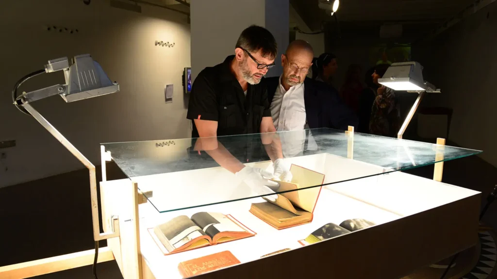 Dos hombres se inclinan sobre una vitrina iluminada; uno, con guantes blancos, sostiene abierto un libro de artista entre otros ejemplares expuestos.