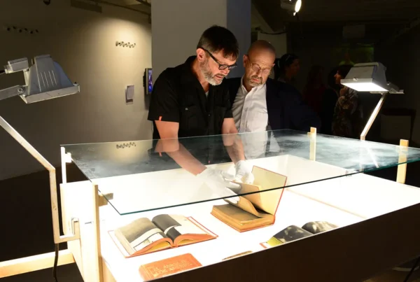 Dos hombres se inclinan sobre una vitrina iluminada; uno, con guantes blancos, sostiene abierto un libro de artista entre otros ejemplares expuestos.