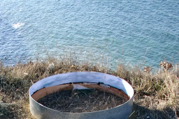 El anillo al filo del acantilado con el mar azul llenando todo el fondo; la pieza recorta un fragmento de paisaje dentro de otro paisaje.