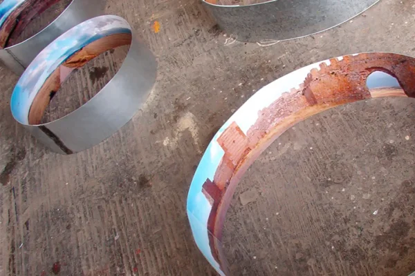 Cuatro anillos vistos en escorzo sobre el suelo de hormigón manchado de pintura; en sus caras interiores se repiten el cielo azul y la muralla de ladrillo.