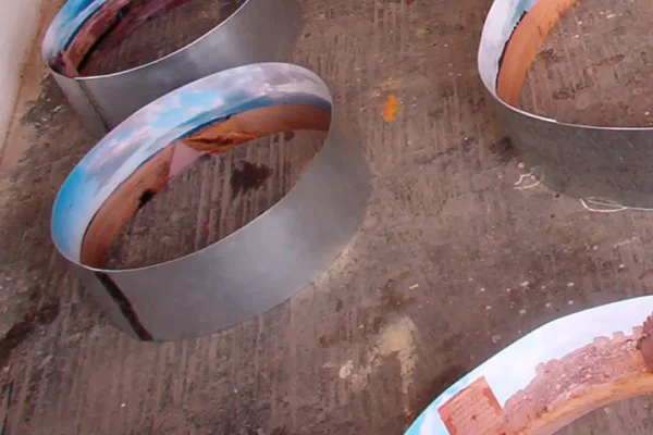 Dos anillos ligeramente inclinados, uno sobre otro; el reflejo curvo del interior mezcla el azul del cielo con el rojo intenso de la tierra.