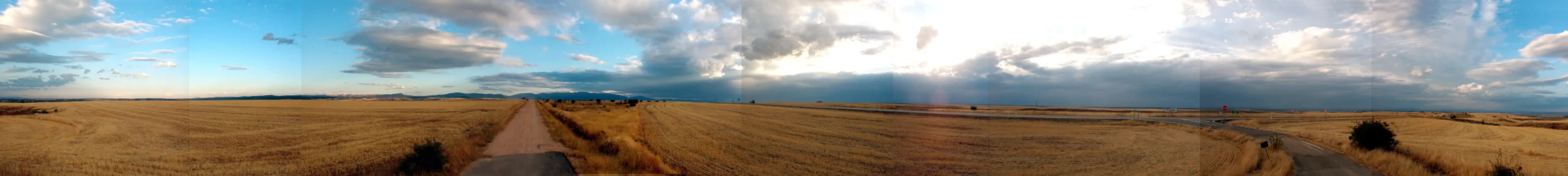 Panorámica de 360°: un camino de tierra atraviesa en línea recta un rastrojo dorado hasta el horizonte, bajo un cielo amplio de nubes dispersas.