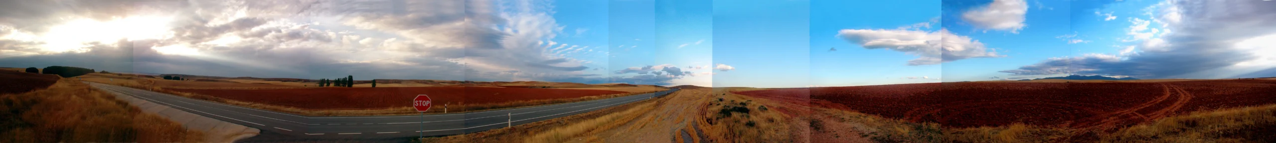 Panorámica de 360°: una carretera cruza campos arados de tierra ocre y roja; una señal de stop en el cruce y rayos de sol atravesando las nubes.