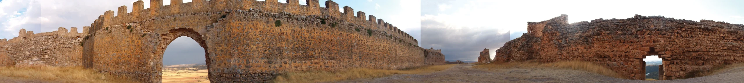 Panorámica de 360° de una muralla almenada de piedra dorada con una puerta de arco apuntado; a la derecha, un tramo del muro ya desmoronado.